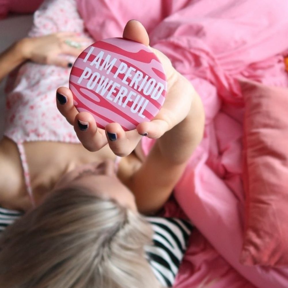 Blonde girl holding up period powerful cosmetic mirror lying down on pink duvet