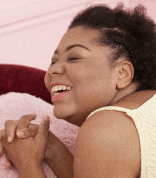 A black female laying on her stomach on a pink bed in a pink room holding her hands. She is smiling and wearing a yellow top with pink shorts