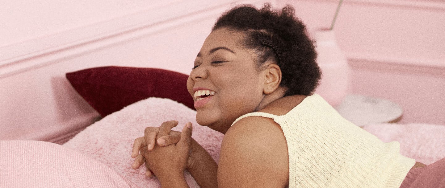 A black female laying on her stomach on a pink bed in a pink room holding her hands. She is smiling and wearing a yellow top with pink shorts