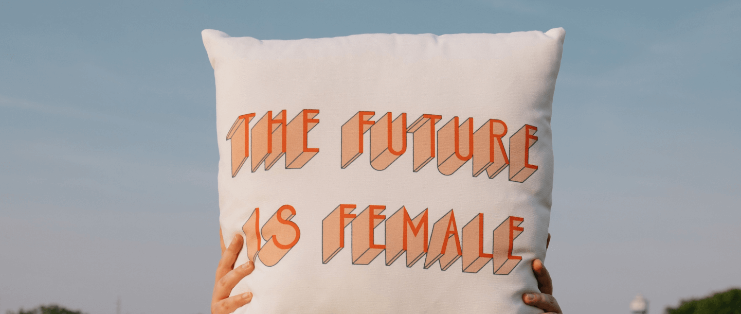 Hands holding up a cushion with the future is female in orange text on it against blue sky background