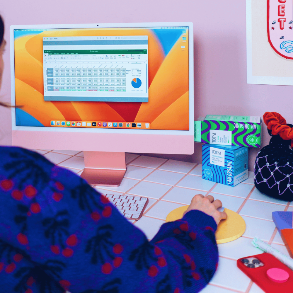 Woman using a computer with period care on the desk as well as other office items like a mouse and mobile phone