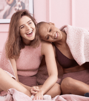 Female PoC in purple bralette and shorts resting her head on the shoulder of a white woman wearing a pink top and purple shorts both sat laughing on a bed in a pink room under a pink blanket to represent girls supporting girls