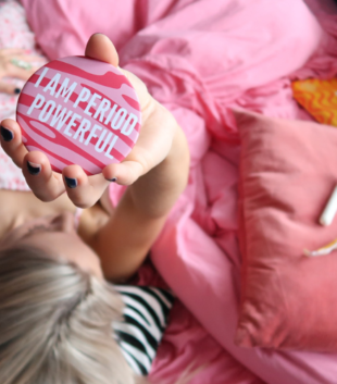 Woman holding period powerful pink mirror with tampons next to her on pillow