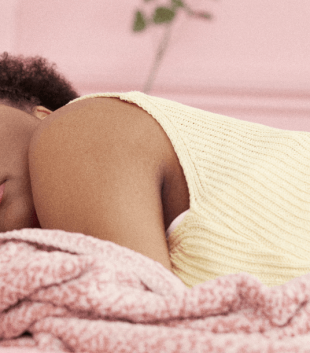 Black female wearing a yellow top and pink shorts laying on chest in a pink bed sleeping on period