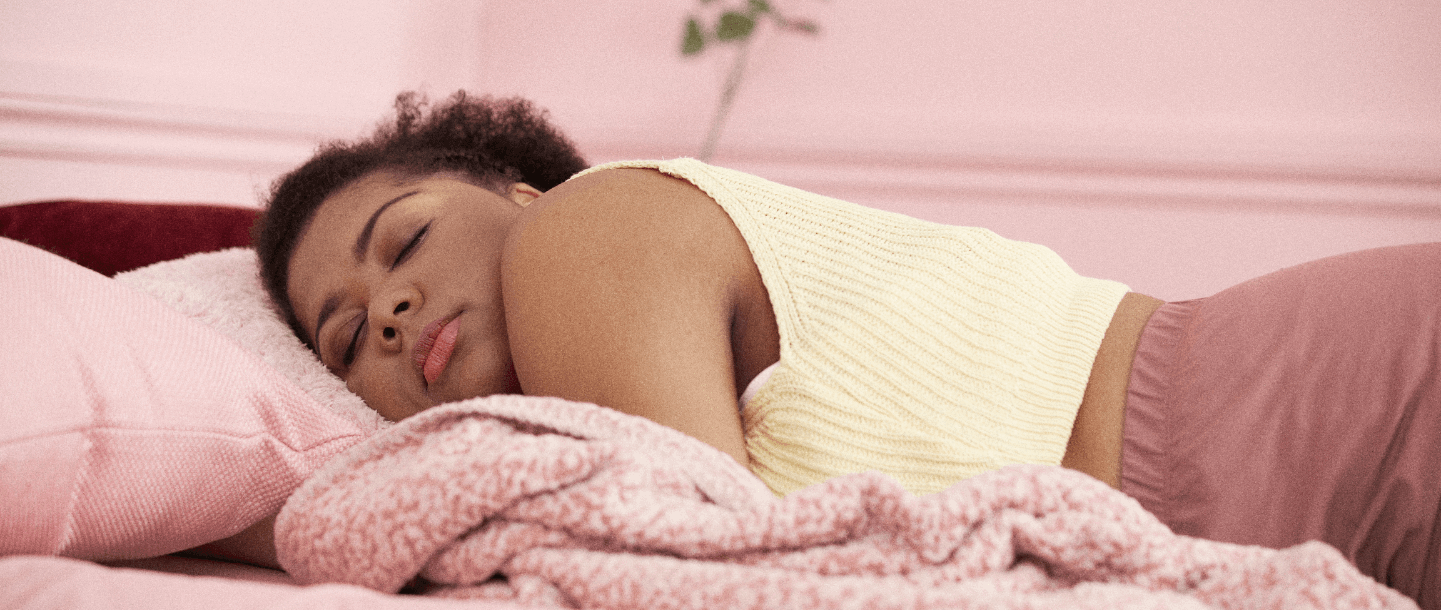 Black female wearing a yellow top and pink shorts laying on chest in a pink bed sleeping on period