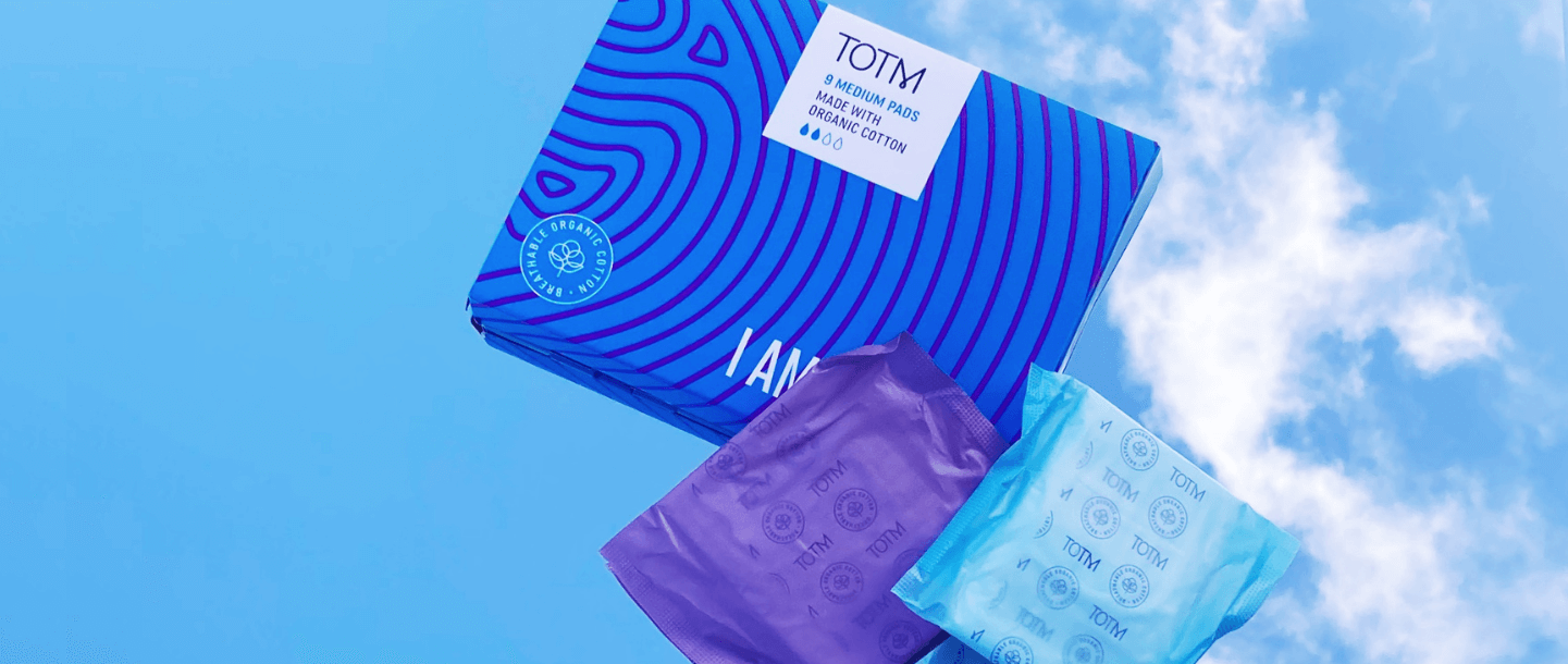 TOTM's medium flow organic cotton pads box next to medium and super pad in their wrapper on a blue background with sky reflection