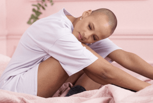 Female PoC in a purple top sitting on a pink bed and leaning forward