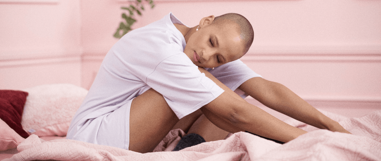 Female PoC in a purple top sitting on a pink bed and leaning forward