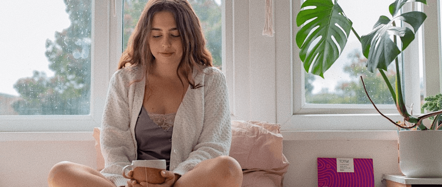 Brunette young female sitting cross legged in front of a window with a herbal tea in her lap to combat flu on her period