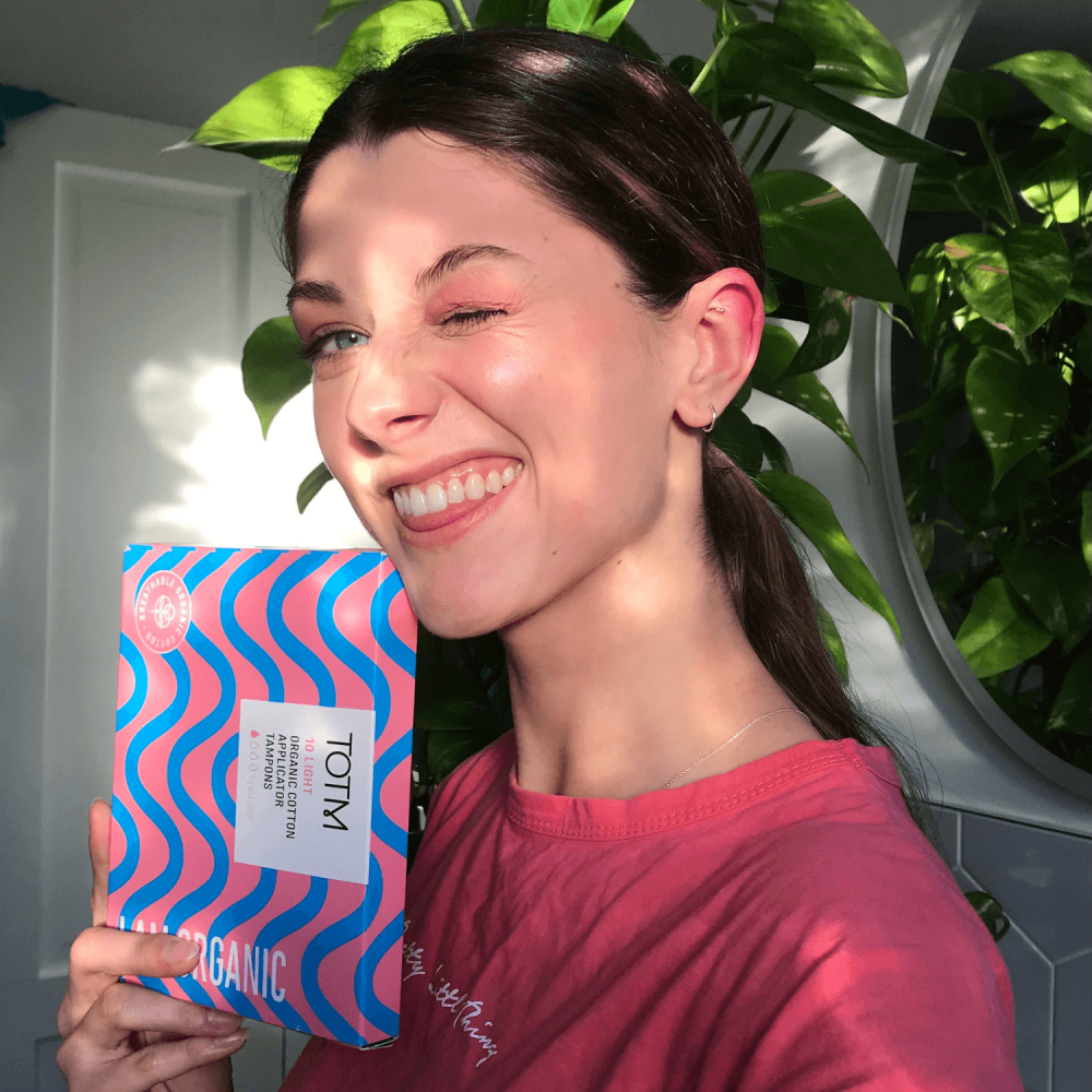 Female winking holding pink and blue TOTM box wearing a red top in front of plant