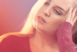 Close-up of a blonde white female tilting her head and resting her fingers on her temple. She is wearing a red top and standing against a purple background