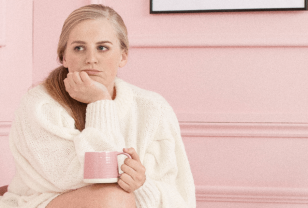 Blonde female is sitting in a pink room wearing a white knitted jumper and is resting her chin on her hand and drinking a herbal tea out of a pink mug which is resting on her knee to combat her period blues