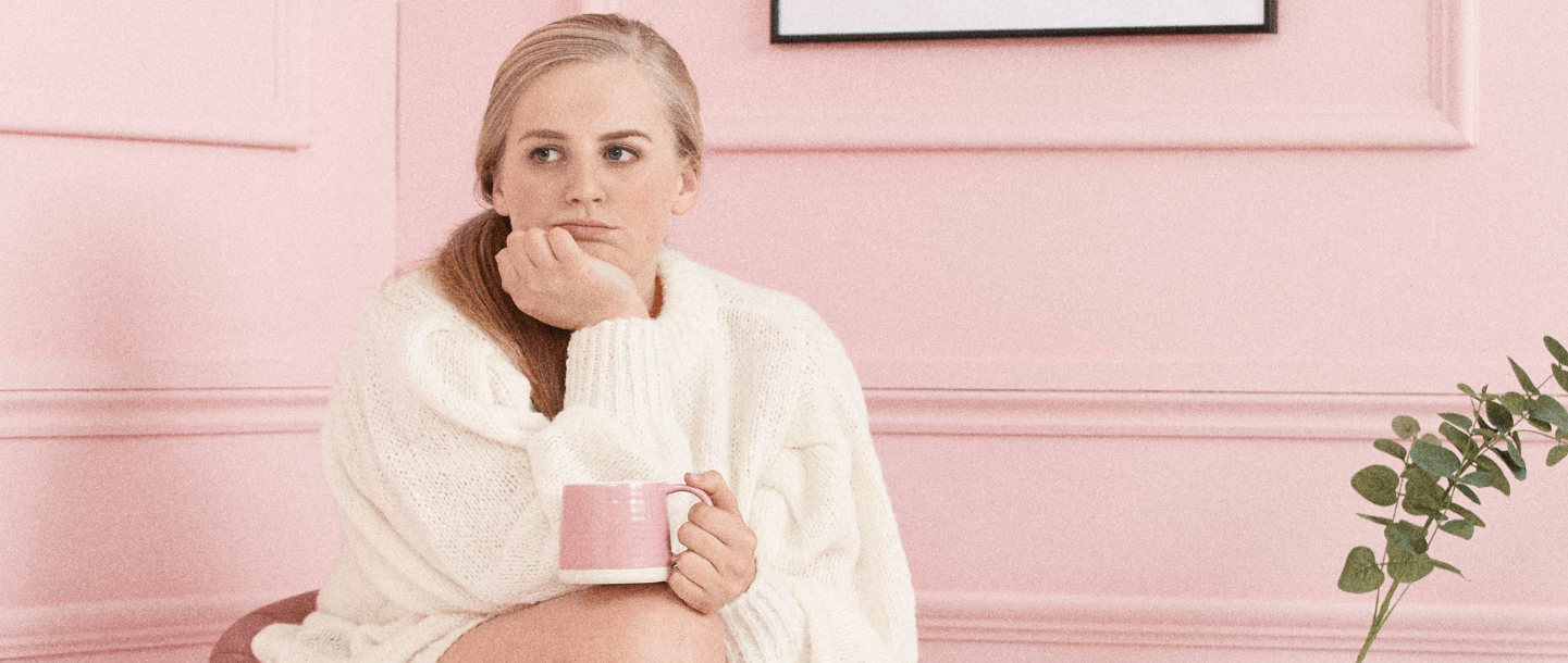 Blonde female is sitting in a pink room wearing a white knitted jumper and is resting her chin on her hand and drinking a herbal tea out of a pink mug which is resting on her knee to combat her period blues