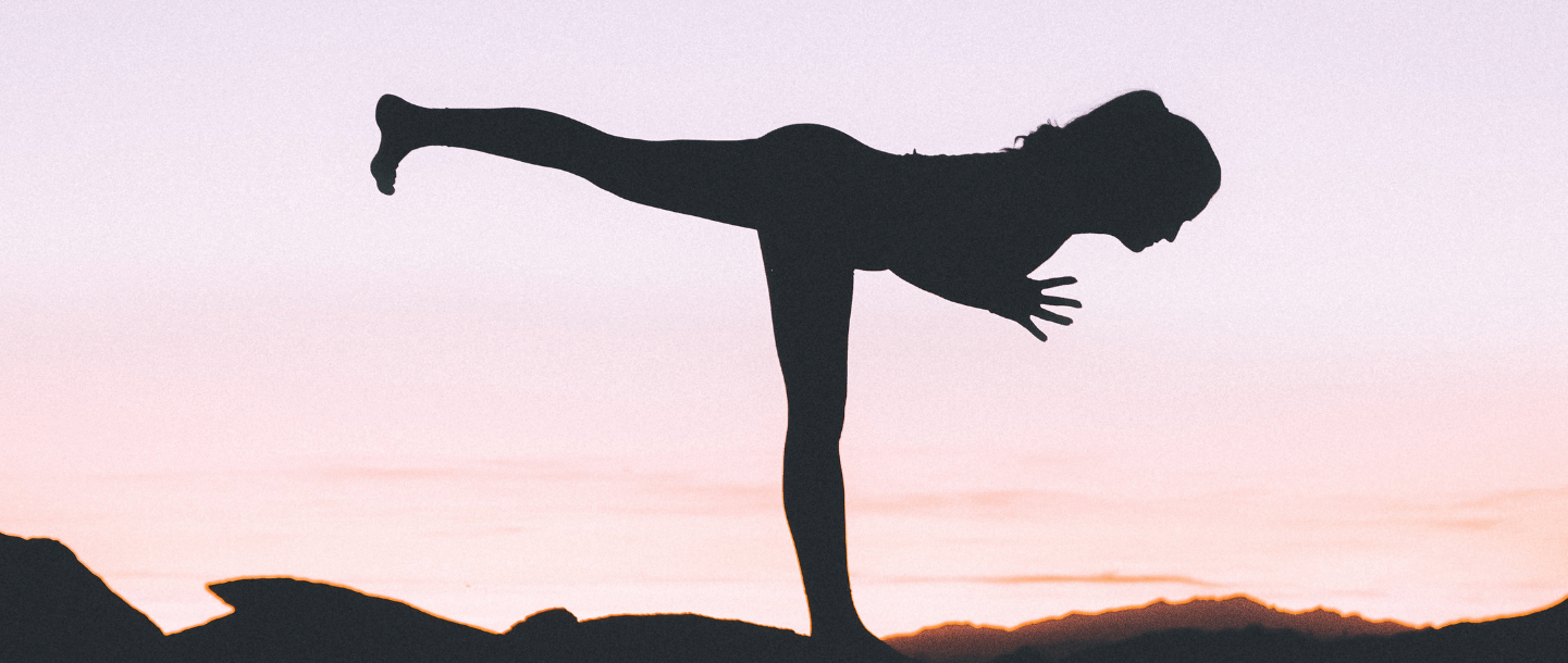 Silhouette of a woman in yoga warrior 3 pose in front of a pastel sky background